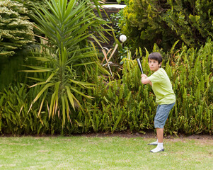 Boy gripping black bat and focusing on white baseball in backyard garden, copy space