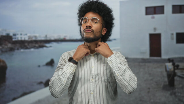 African american man adjusting shirt on seaside promenade with ocean backdrop and modern glasses, displaying casual outdoor fashion scene near water and rocks.