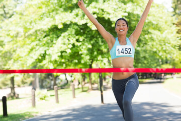 Asian woman running through red finish line tape on park path with turquoise top bib 1452