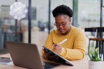 Focused african american businesswoman is taking notes in her notebook during a business meeting, utilizing a laptop and tablet in a contemporary office setting