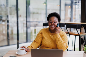Smiling businesswoman talking on mobile phone and working on laptop while enjoying a cup of tea in a modern cafe, demonstrating efficient multitasking and work-life balance