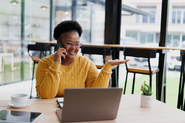 Young black female freelancer is talking on phone and gesturing while working on laptop in a cafe, enjoying a cup of coffee and using a tablet