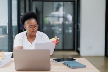 Young office worker concentrating on paperwork, holding documents with colorful sticky notes, sitting at desk with laptop, tablet, and notebook in a modern office environment