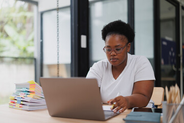 Fototapeta premium Focused african american businesswoman is working on her laptop and taking notes during an online meeting in a modern office, demonstrating professionalism and dedication to her work