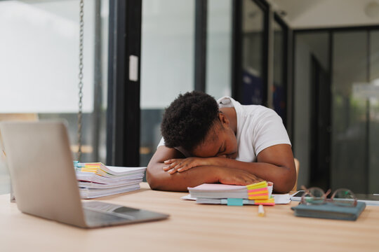 Young African American student sleeping on her desk, surrounded by paperwork and a laptop, showing signs of exhaustion after long hours of studying and working hard on assignments