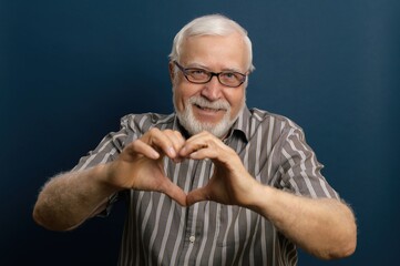 Elderly man makes heart shape with hands against a blue background while smiling warmly