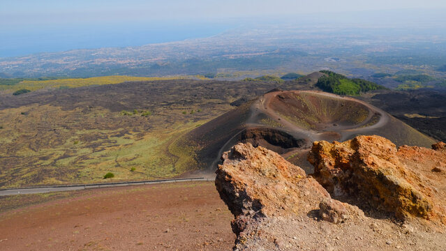 Rugged volcanic landscape with large, rust-colored rock, and cinder cone and volcanic crater. Powerful beauty of volcanic region, unique environment of Mount Etna. Travel, nature, geology.