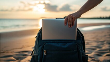 Fototapeta premium A person places a laptop into a backpack on a sandy beach at sunset.