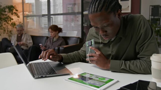 Young African American mansitting at office desk usig laptop and digital table and learning how to use slar charger device