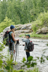 Woman standing with hiking poles near river talking to boy drinking water, both wearing backpacks, surrounded by lush green forest and rocky landscape
