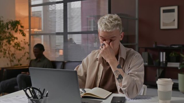 Tired young Caucasian man aking off his eyeglasses ad rubbing nose bridge whil working on laptop in offie