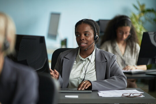 Black woman wearing headset working at computer in modern office, engaging in customer service or technical support, colleagues working in background