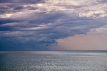 Expansive view of baltic sea with textured clouds looming overhead, capturing dramatic lighting. Calm waters reflect subtle pastel hues. Atmospheric perspective creates serene and contemplative mood