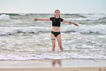 Young female with red hair performs yoga-like stance in baltic sea. Overcast sky and grey waves create serene, energetic atmosphere. Water splashes around her legs