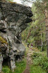 Family hiking along rocky forest trail near large stone cliff, carrying backpacks and trekking poles, surrounded by dense green trees and rugged terrain