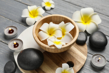 Beautiful spa composition with tray, stones and bowl with plumeria flowers on grey wooden background, closeup