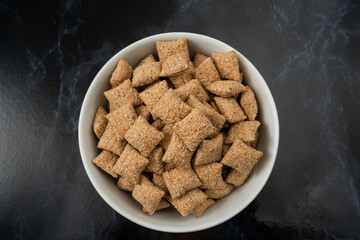 A bowl filled with small, square-shaped, crunchy cereal pieces sits on a sleek black marble surface, perfect for a nutritious breakfast or snack option any time of day.