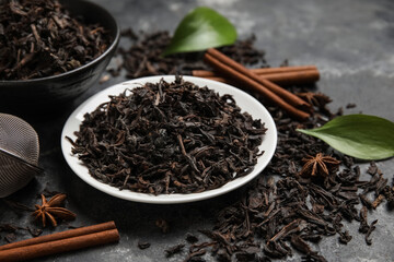 Plate with dry black tea leaves and cinnamon on dark background, closeup