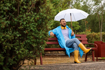 Fototapeta premium Young man with blue raincoat and umbrella sitting on bench in park