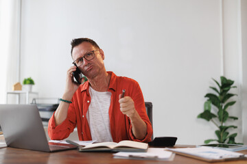 Stressed mature entrepreneur engaging in an intense phone conversation, holding a pen while working...