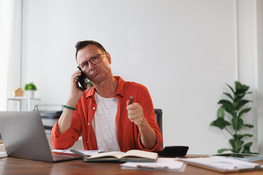 Stressed mature entrepreneur engaging in an intense phone conversation, holding a pen while working on a laptop and reviewing documents at a home office desk
