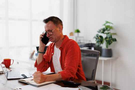 Businessman getting angry and stressed while working at the computer and receiving bad news on the phone, he is holding his fist and looking worried