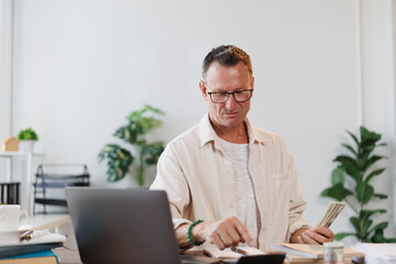 Obraz premium Focused mature entrepreneur calculating domestic expenses, managing personal finances, counting cash money and using calculator, working from home