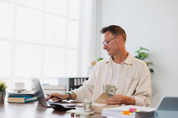 Obraz premium Businessman holding and counting american dollars while working on a laptop in his bright office, he is managing his finances and investments