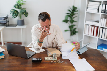 Overwhelmed businessman experiencing financial problems, managing budget and calculating bills, sitting at his desk with laptop, calculator, money and paperwork, showing frustration and stress