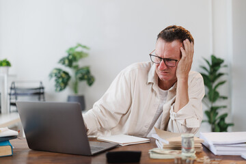 Businessman with head in hand looking stressed and having financial problems while working on laptop at home office desk with notepad, calculator, money and paperwork