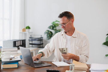 Obraz premium Mature entrepreneur working with laptop and counting american dollars while sitting at wooden table at home office, managing his business finances