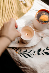 Hand reaching toward a burning candle near a cup of hot tea with citrus slice. Cozy intimate moment with warm rustic home atmosphere.
