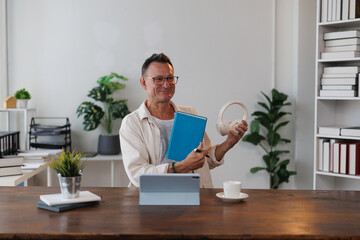 Smiling mature professor showing book and headphones while sitting at desk with digital tablet, cup of tea and notebooks during online class at home
