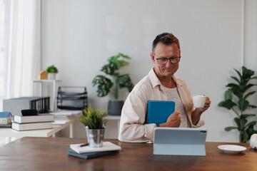 Senior architect enjoying a coffee break, checking his tablet and holding a book in his modern office, with plants and scale models on his desk