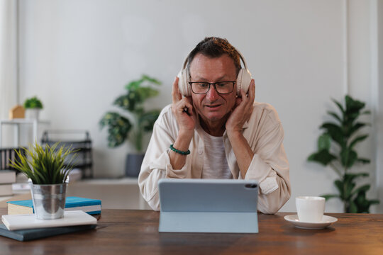 Mature professor with eyeglasses and headphones attending online course using tablet and enjoying music at home, plants and cup of coffee on desk, concept of online education and learning