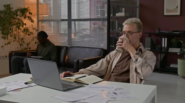 Young gen Z male specialis looking at charts, drinkig coffee and working on latop in modern office
