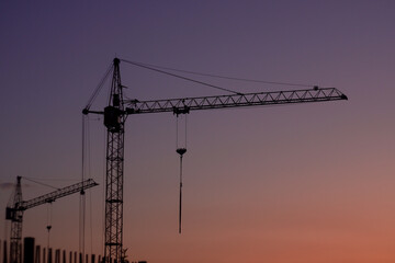 Two construction cranes stand against a gradient sky transitioning from purple to orange. The cranes are silhouetted, with a clear view of their structure and cables.