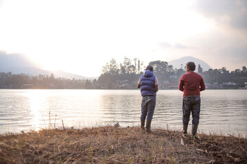 Fototapeta premium Two Men Standing on a Grassy Bank at Sunrise Fishing in a Lake With a Scenic View of Mountains