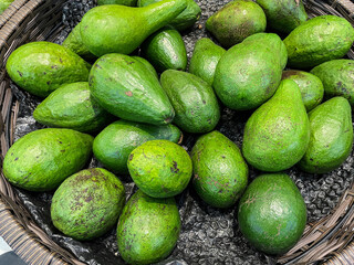 A bountiful display of fresh green avocados piled in a dark woven basket. Their varied shapes and vibrant color highlight their natural, healthy appeal at a market stall