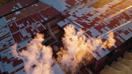 AI-generated of aerial of rusted metal rooftops emitting steam at an industrial facility at sunset - Powered by Adobe