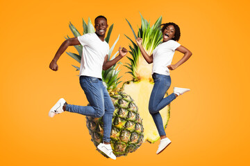 African American cheerful couple dressed in white tees and jeans jumps with enthusiasm against an orange backdrop featuring large pineapples. Their laughter embodies a fun, tropical vibe.