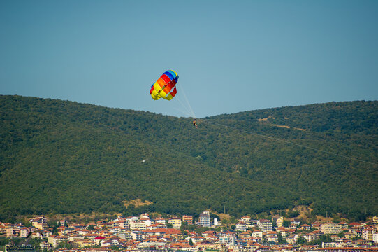 A parasailer floats above a scenic town and forested hillside, tethered to a vivid red, yellow, and blue parachute in Bu.garia