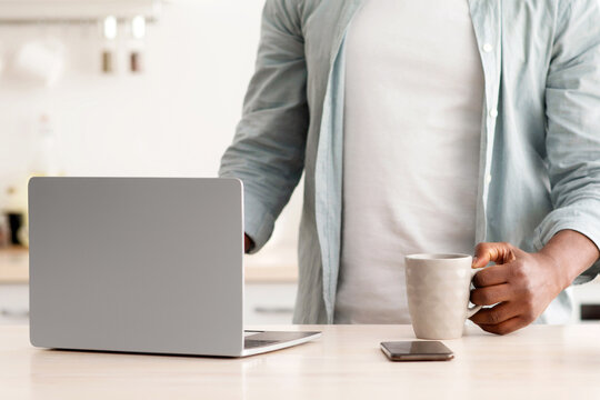 Morning routine concept. Unrecognizable african american man using laptop and drinking hot beverage in kitchen, browsing social media or reading online news, crop, free space