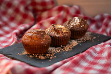 Homemade muffins on slate board over red checkered cloth, warm rustic kitchen setting, close-up food photography.
