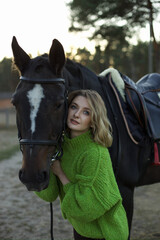 A young woman in a green sweater leans affectionately against her horse in a calm equestrian setting. The background features trees and soft evening light.