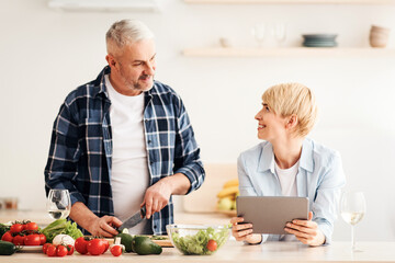 Cooking in kitchen together, searching recipe in internet, watch online blog. Happy middle aged man cuts vegetables for salad, woman with tablet look food lesson in kitchen interior, empty space