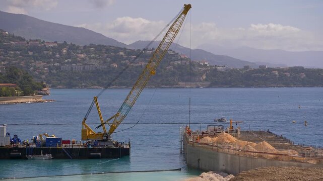 A large yellow crane operates from a barge next to an offshore construction platform, where piles of sand and building materials are visible - Monaco 7 May 2019