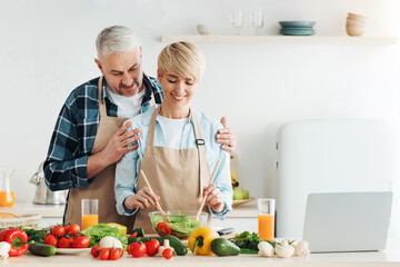 Enjoy of cooking healthy food together, modern blog and love. Happy middle aged european man hugs smiling wife making salad in modern kitchen interior with bright fresh vegetables on table with laptop