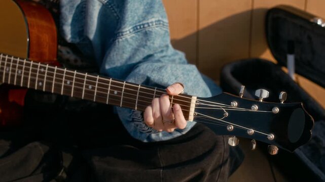 Medium closeup of unrecognizable female busker sitting outdoors, playing acoustic guitar