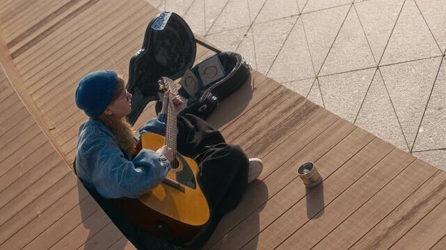 High angle view of young ault female street musiciansitting on wooden bench inpublic space, playing guitr and singing